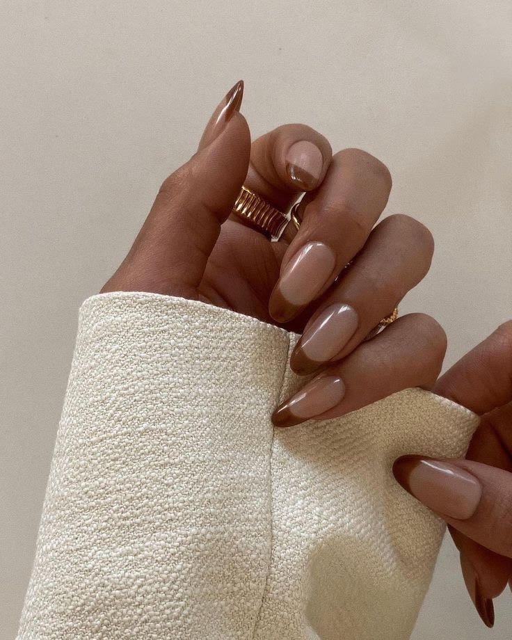A hand with polished, almond-shaped nails in shades of nude and brown—perfect autumn nail colors—adorned with gold rings, holds a textured, cream-colored fabric.